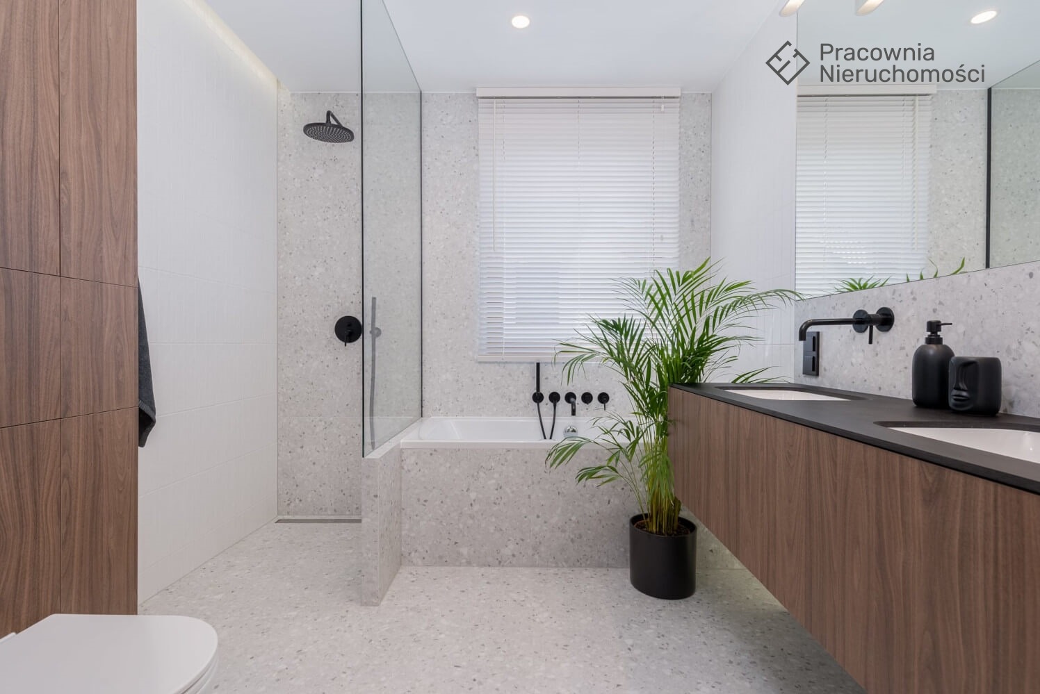 Nowoczesna łazienka z kabiną prysznicową typu walk-in, wanną obudowaną płytkami lastryko, podwójną umywalką z szafką z forniru orzechowego i czarną armaturą, uzupełniona zieloną rośliną doniczkową.