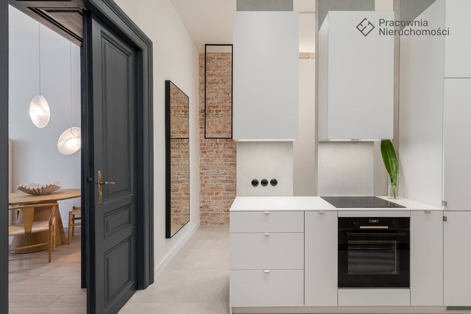 Nowoczesna kuchnia z białymi szafkami, czarnym piekarnikiem i płytą indukcyjną, widok na jadalnię przez otwarte, ciemnoszare drzwi, ceglana ściana w tle.