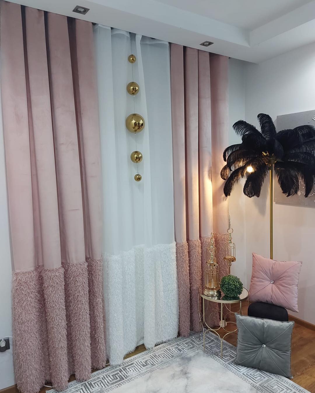 Eleganckie, długie zasłony w odcieniach pudrowego różu i bieli z dekoracyjnym futerkiem u dołu, ozdobione złotymi kulami zawieszonymi na żyłce, uzupełnione lampą w kształcie palmy i stolikiem...