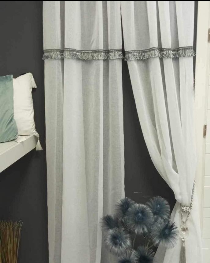 Delikatne, półprzezroczyste zasłony w odcieniach bieli z dekoracyjnym srebrnym wykończeniem i frędzlami, upięte ozdobnym sznurem z pomponami, na tle ciemnoszarej ściany z półką i dekoracją kwiatową.