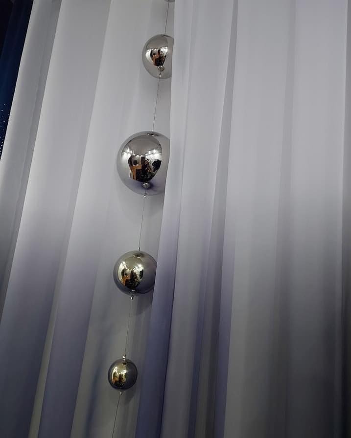 Delikatna, biała zasłona z ombre przechodzącym w szarość, ozdobiona pionowym sznurem z czterema srebrnymi, odbijającymi światło kulami.