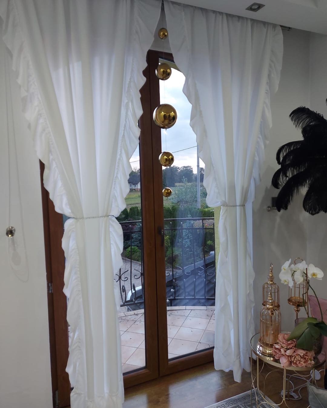 Eleganckie, białe zasłony z falbanami, upięte srebrnymi pasami, wiszące w oknie balkonowym, z dekoracyjnymi złotymi kulami na sznurkach. Na stoliku obok dekoracje w stylu glamour: kwiaty i złota...