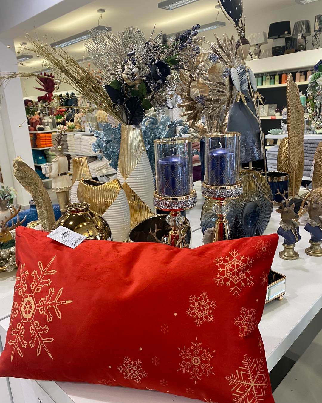 Czerwona poduszka dekoracyjna z motywem złotych płatków śniegu na tle ekspozycji sklepu z artykułami wyposażenia wnętrz, w tym wazony, świeczniki i figurki.