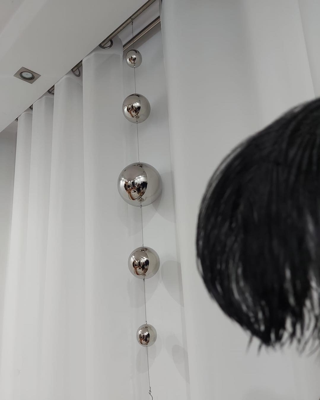 Biała, gładka zasłona z dekoracyjnymi, srebrnymi kulami zwisającymi na żyłkach, częściowo zasłonięta czarnym pióropuszem po prawej stronie.