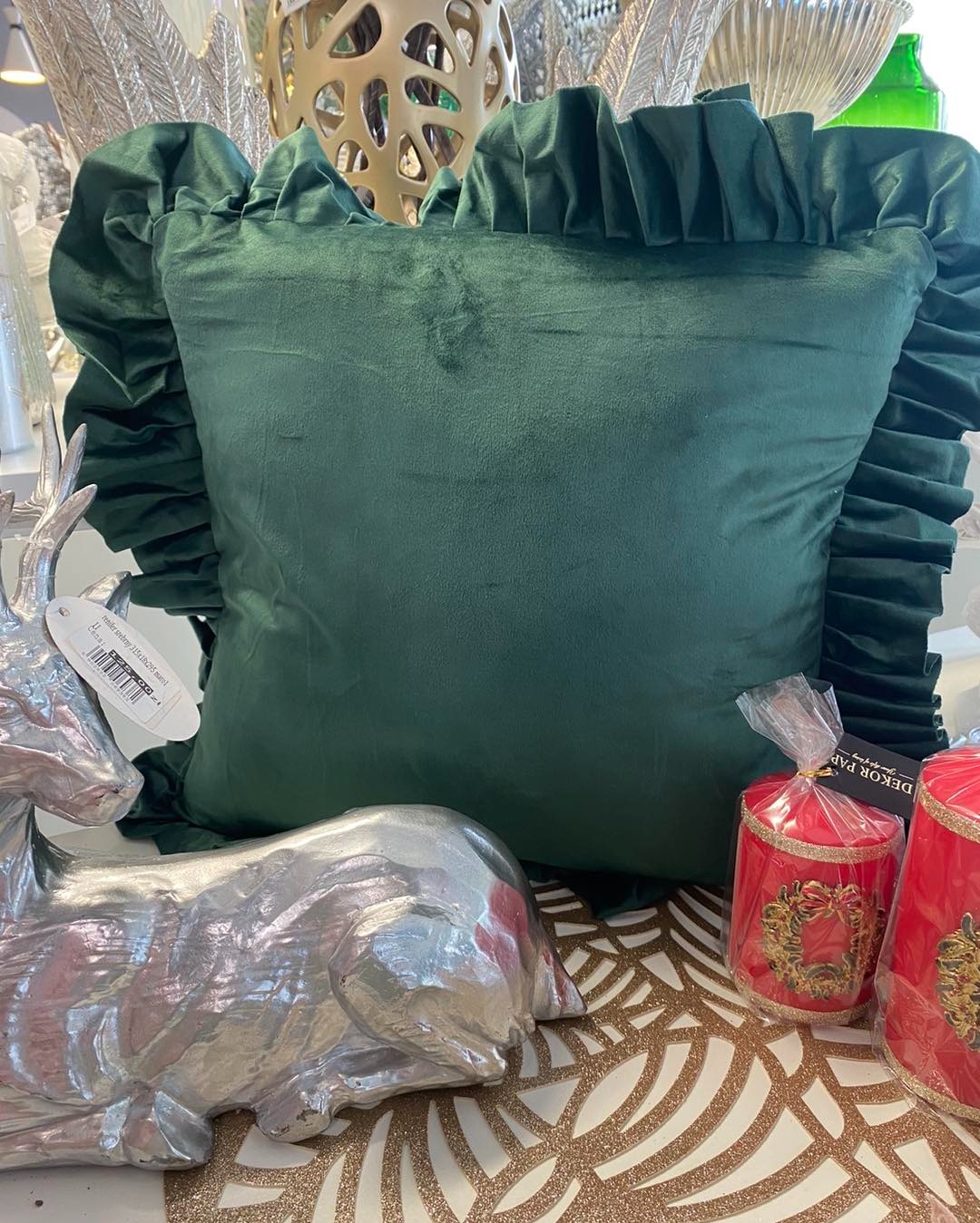 Ciemnozielona, kwadratowa poduszka z falbanami, ustawiona na stole obok srebrnej figurki renifera i dwóch czerwonych świec z dekoracjami świątecznymi.