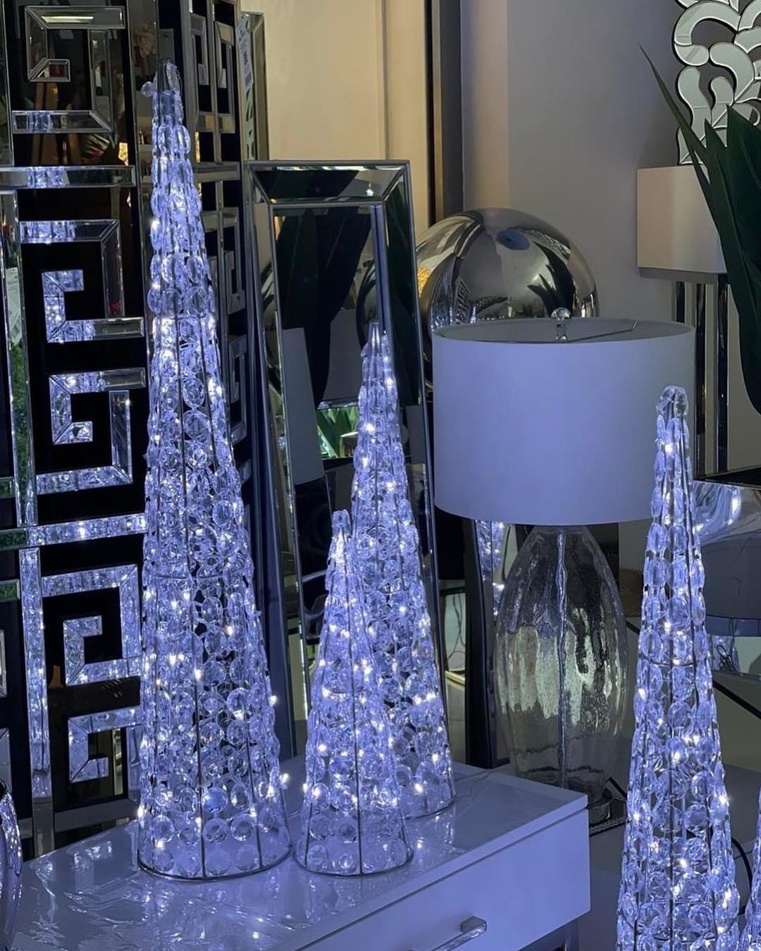 Dekoracyjne, kryształowe stożki z oświetleniem LED, ustawione na błyszczącej komodzie, odbijające się w lustrzanej ścianie z geometrycznym wzorem.