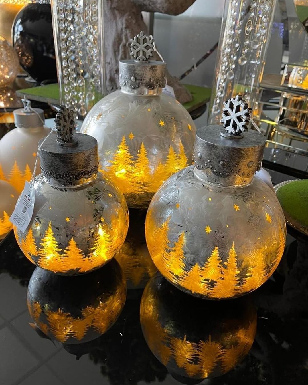 Trzy szklane bombki choinkowe z podświetlanymi, złotymi motywami choinek i srebrnymi płatkami śniegu na wierzchu, odbijające się w błyszczącej powierzchni.