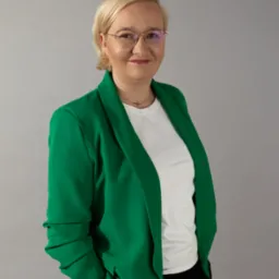 Portret uśmiechniętej kobiety w okularach, blond włosach i zielonej marynarce na szarym tle.