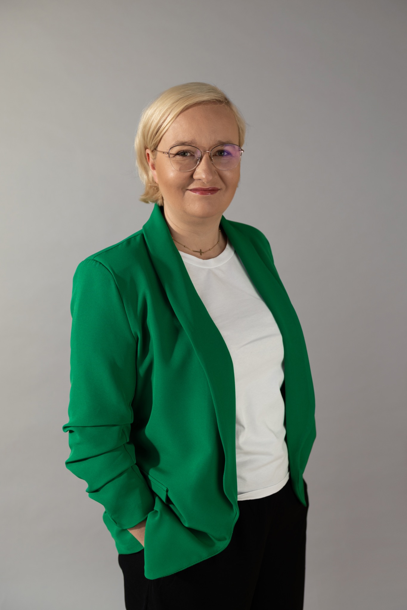 Portret uśmiechniętej kobiety w okularach, blond włosach i zielonej marynarce na szarym tle.