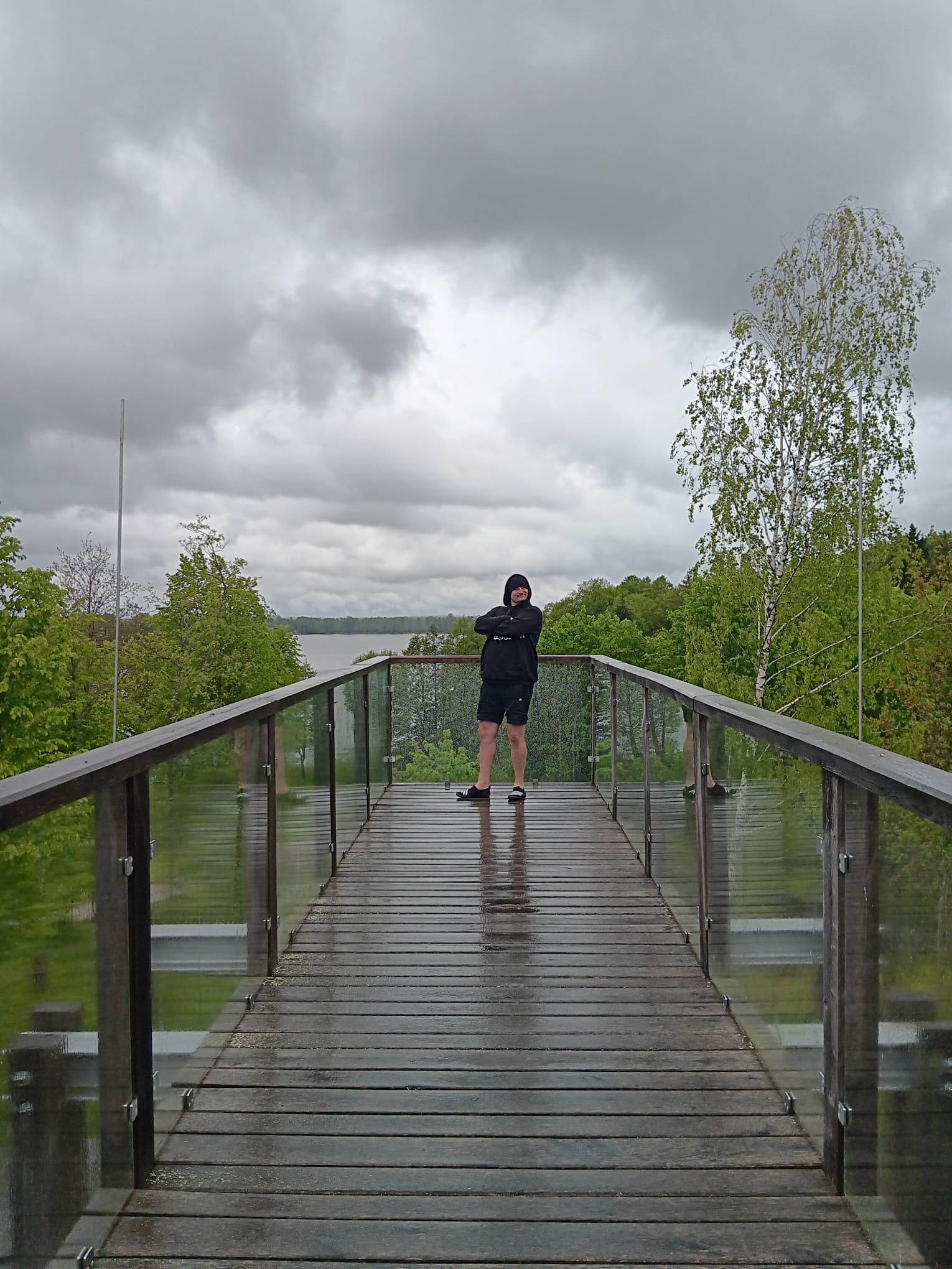 Mężczyzna w czarnej bluzie stoi na drewnianym pomoście z szklanymi barierkami, w tle jezioro i pochmurne niebo. Widok na krajobraz z platformy widokowej.
