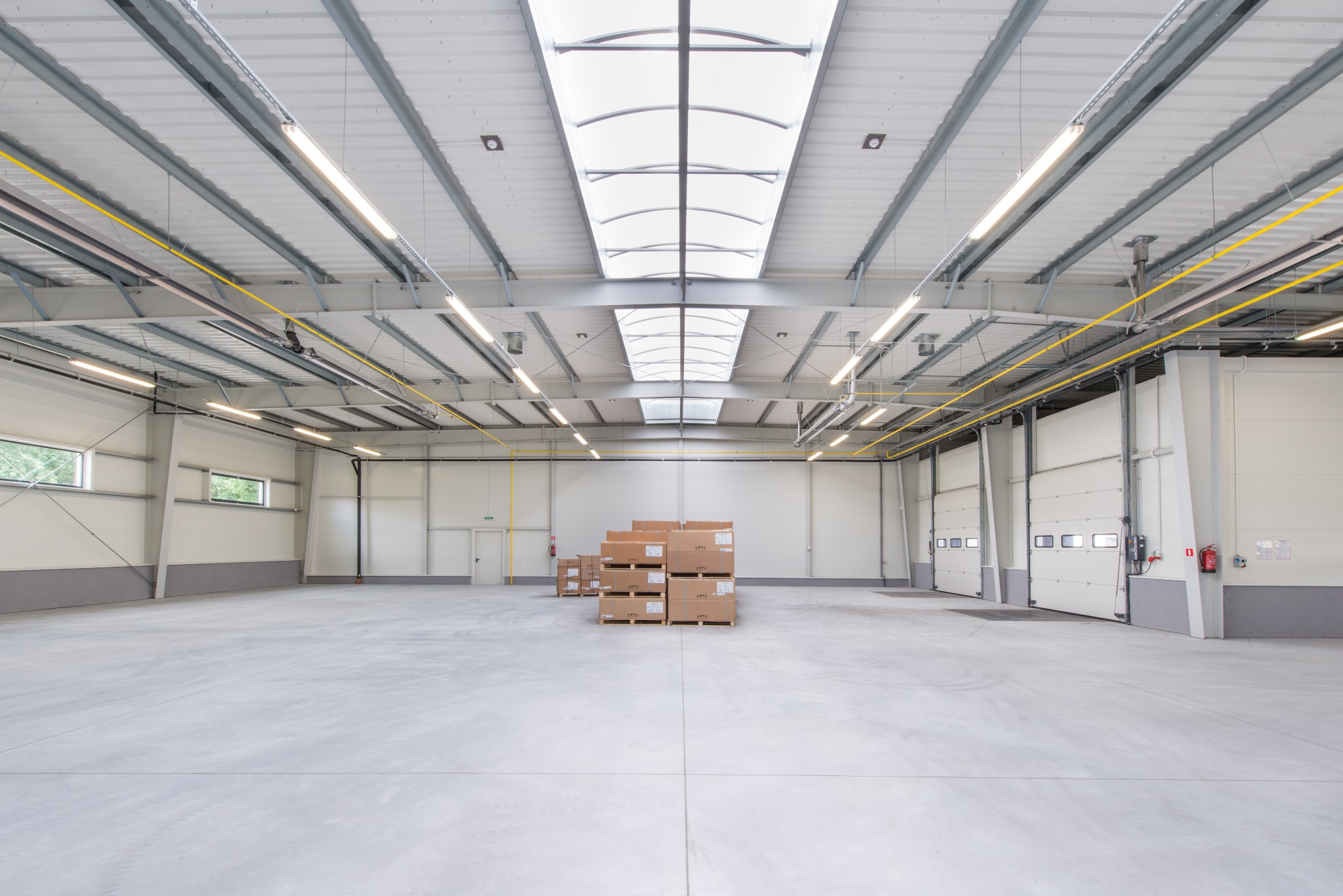 Wnętrze nowo wybudowanej hali magazynowej z betonową posadzką, widoczne stalowe elementy konstrukcyjne pod dachem i świetliki, palety z kartonami w centralnej części, oraz bramy garażowe w tle.