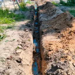 Wykop pod przyłącze elektryczne z widocznymi niebieskimi rurami osłonowymi kabli, w tle częściowo widoczny mini-koparka i budynek w trakcie budowy.