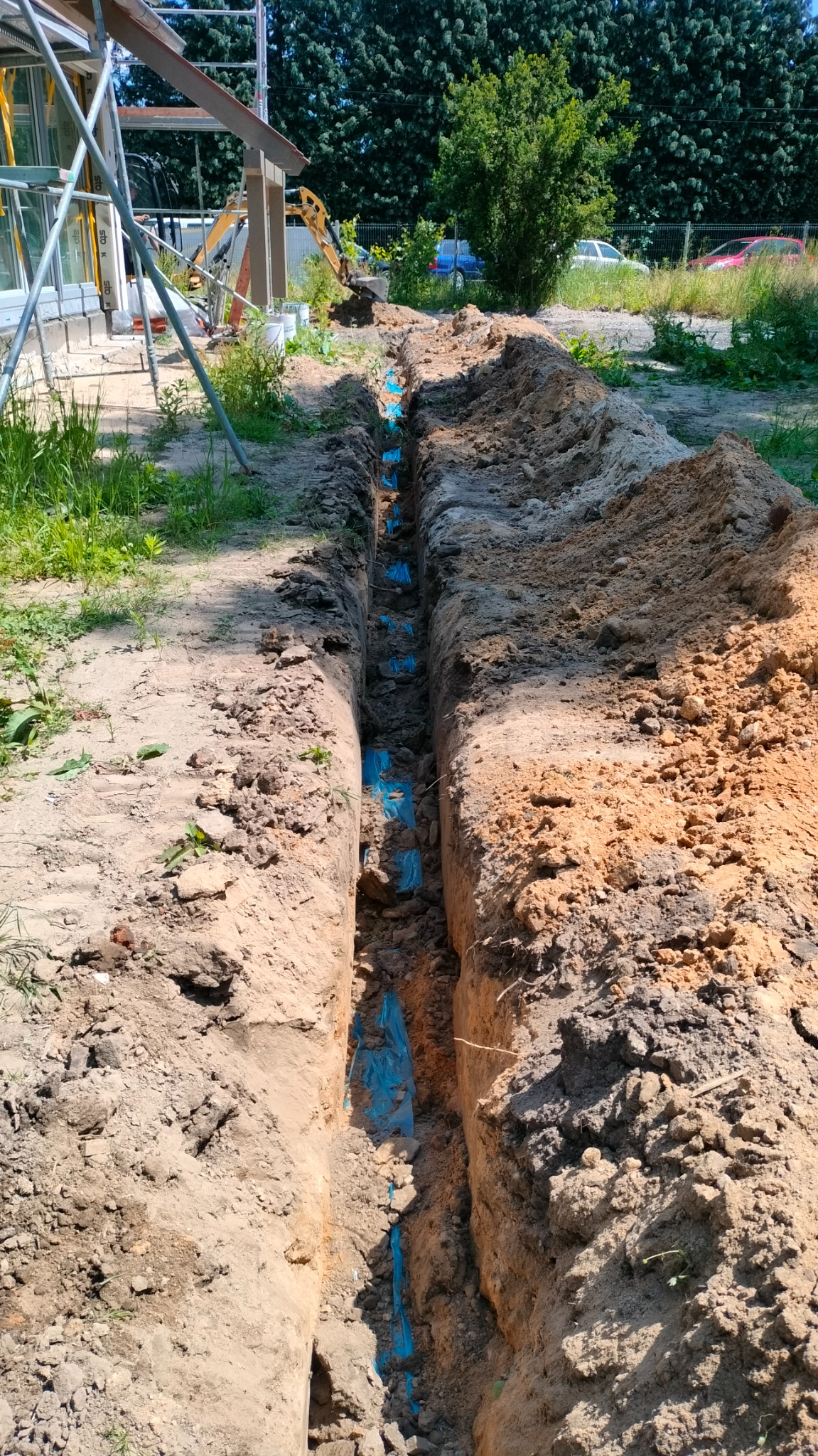 Wykop pod przyłącze elektryczne z widocznymi niebieskimi rurami osłonowymi kabli, w tle częściowo widoczny mini-koparka i budynek w trakcie budowy.