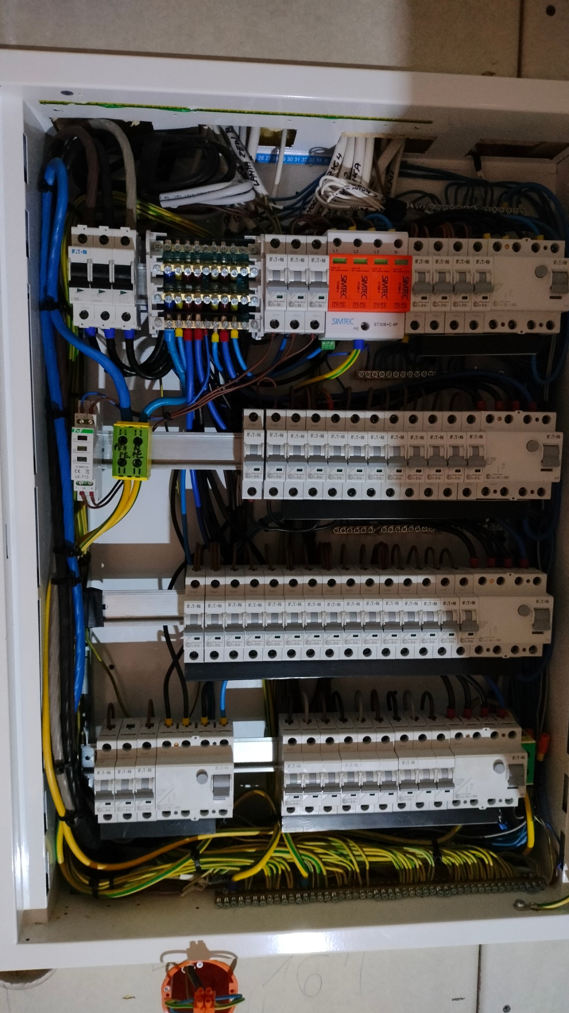 Otwarta skrzynka rozdzielcza z widocznymi bezpiecznikami, przewodami i listwami zaciskowymi, z elementami oznaczonymi kolorami i etykietami, zamontowana na ścianie.