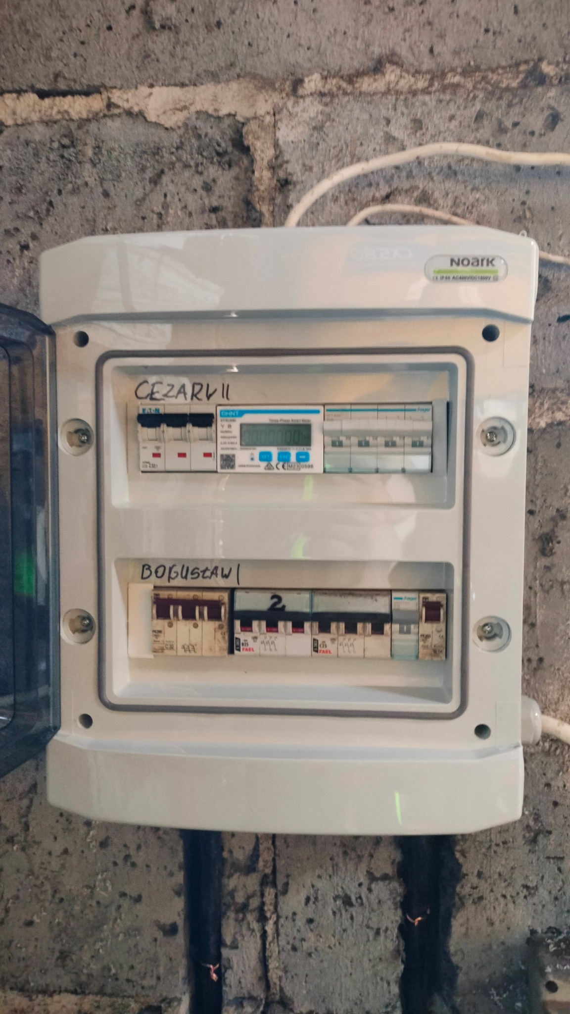 Otwarta skrzynka elektryczna z bezpiecznikami i licznikiem energii, zamontowana na surowej, betonowej ścianie. Widoczne napisy 'Cezary II' i 'Bogusław' nad rzędami bezpieczników. W prawym górnym...
