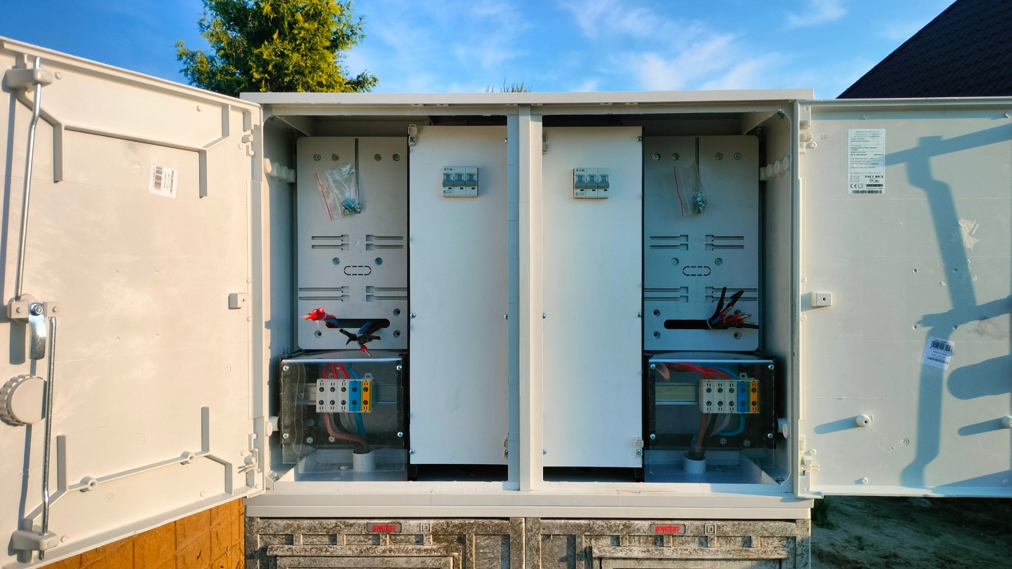 Otwarta, podwójna skrzynka elektryczna z widocznymi elementami instalacji wewnętrznej, w tym przewodami, bezpiecznikami i listwami zaciskowymi, zamontowana na zewnątrz budynku w słoneczny dzień.