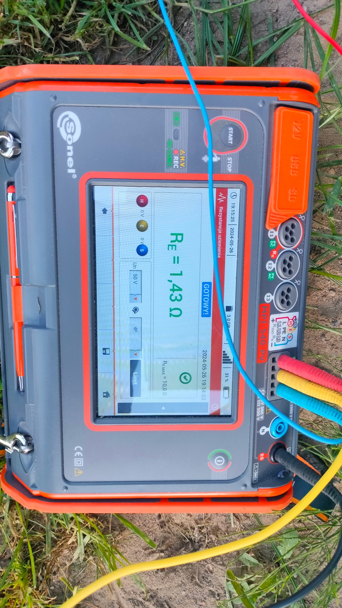 Miernik Sonel MPI-540-PV na trawie, wyświetlający wynik pomiaru rezystancji uziemienia Re=1,43 Ω, z podłączonymi przewodami pomiarowymi w kolorach niebieskim, czerwonym i żółtym.