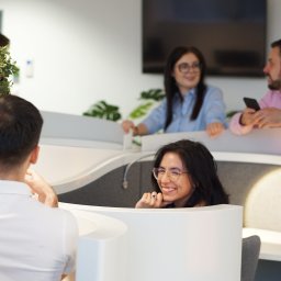 DKLAW Paulina Jachna Kancelaria Radcy Prawnego - Współczesna przestrzeń biurowa z otwartymi kabinami, w których pracują uśmiechnięci ludzie, widoczna dekoracyjna roślina w doniczce.