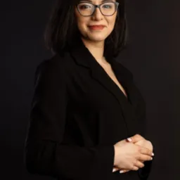 Portret uśmiechniętej kobiety w czarnej marynarce i okularach na ciemnym tle, dłonie splecione przed sobą.