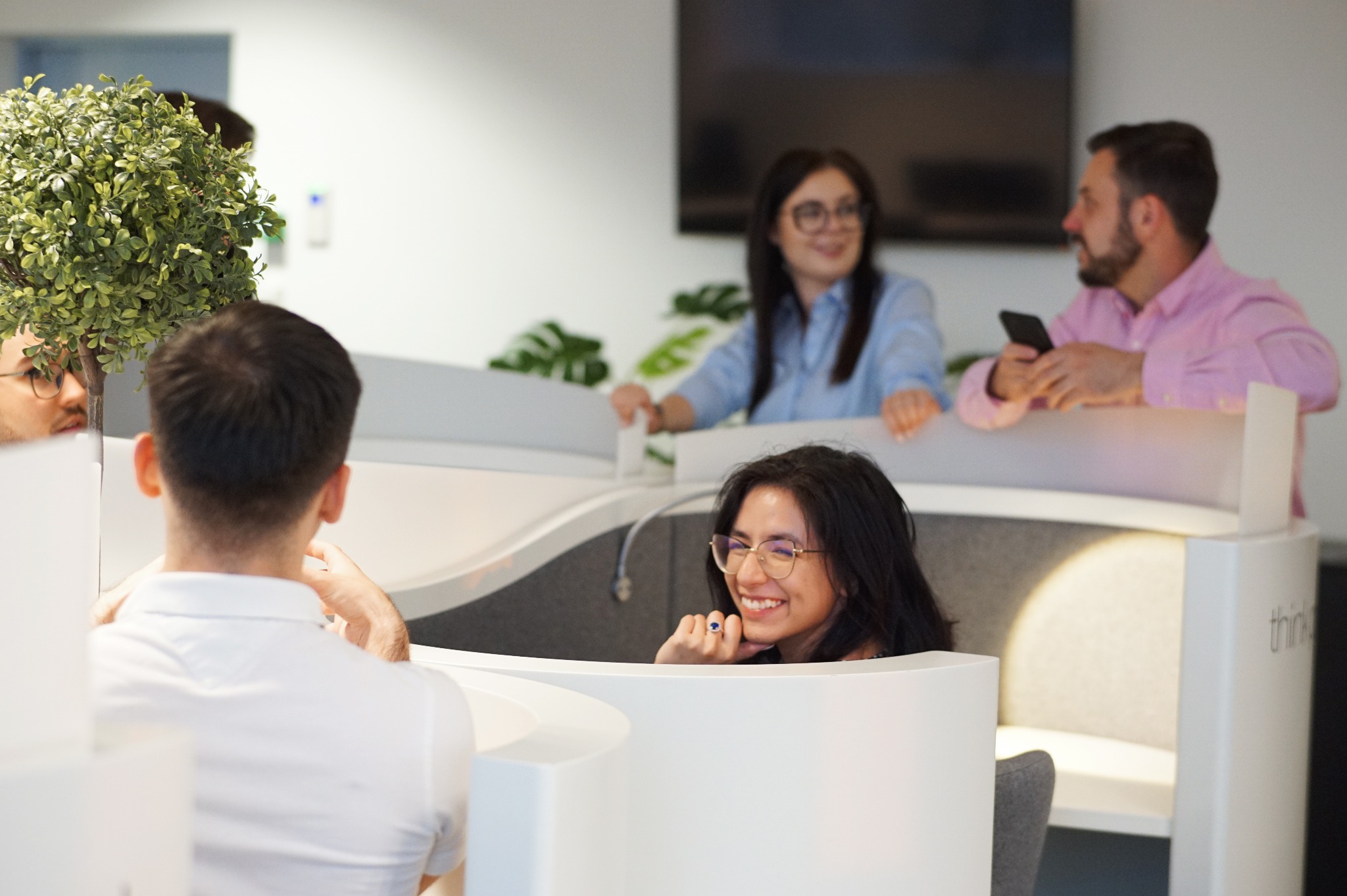 Współczesna przestrzeń biurowa z otwartymi kabinami, w których pracują uśmiechnięci ludzie, widoczna dekoracyjna roślina w doniczce.