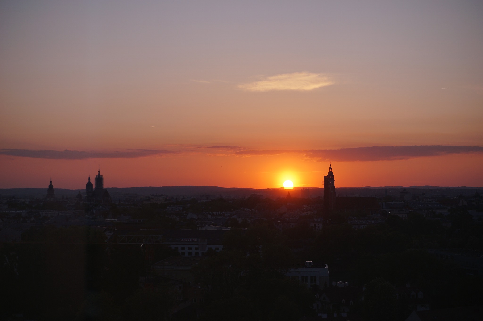Panorama miasta o zmierzchu z widocznymi wieżami kościołów i ratusza na tle zachodzącego słońca.