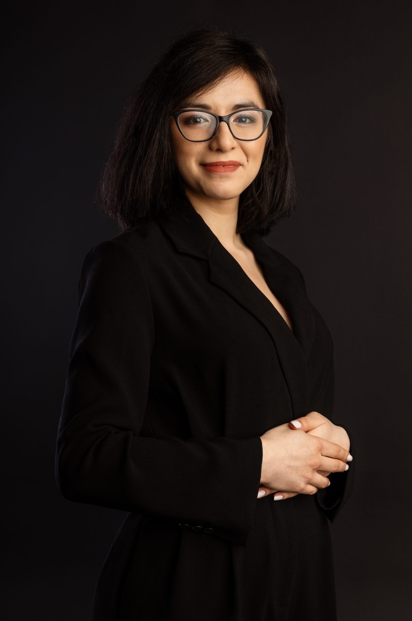 Portret uśmiechniętej kobiety w czarnej marynarce i okularach na ciemnym tle, dłonie splecione przed sobą.