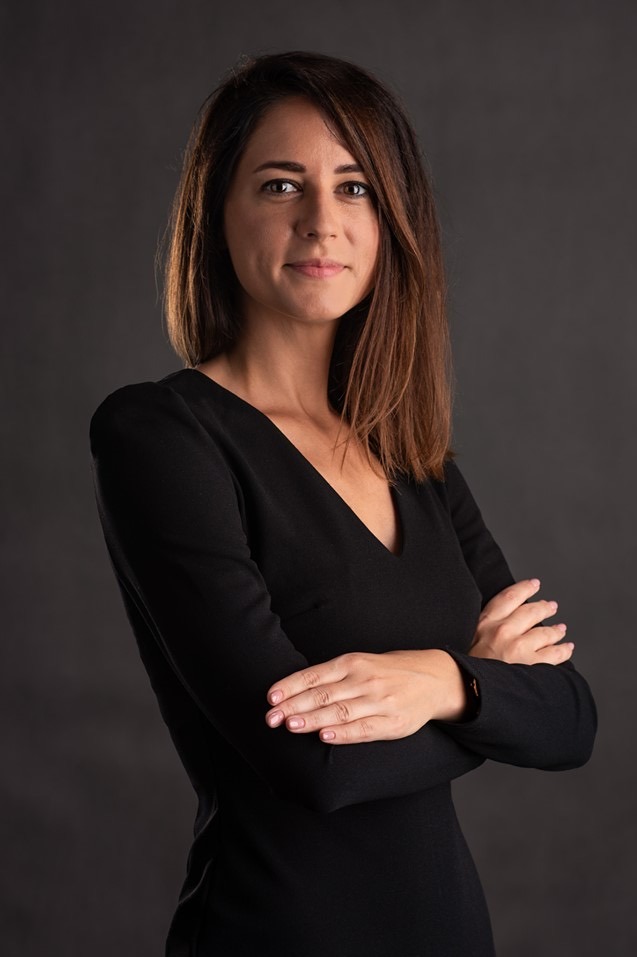 Portret kobiety z brązowymi włosami do ramion, ubranej w czarną sukienkę z rękawami, z założonymi rękami na tle ciemnoszarego gradientu.