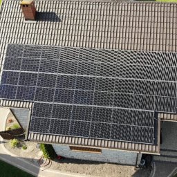 Chrzanowski oze - Widok z góry na dom z brązowym dachem krytym dachówką i zainstalowanymi panelami słonecznymi, obok komina. Układ paneli dopasowany do kształtu dachu, widoczne ścieżki i ogród.