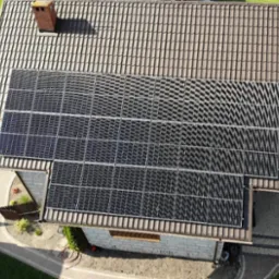 Widok z góry na dom z brązowym dachem krytym dachówką i zainstalowanymi panelami słonecznymi, obok komina. Układ paneli dopasowany do kształtu dachu, widoczne ścieżki i ogród.