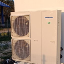 Chrzanowski oze - Zewnętrzna jednostka pompy ciepła Panasonic zamontowana na metalowej konstrukcji na betonowej podstawie, z widocznym fragmentem budynku w tle.