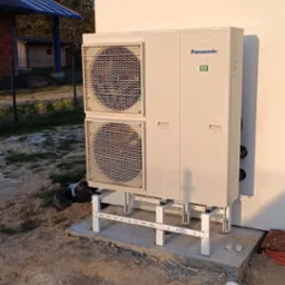 Zewnętrzna jednostka pompy ciepła Panasonic R32 zamontowana na metalowym stelażu przy ścianie budynku, widoczny fragment dachu z panelami solarnymi w tle.