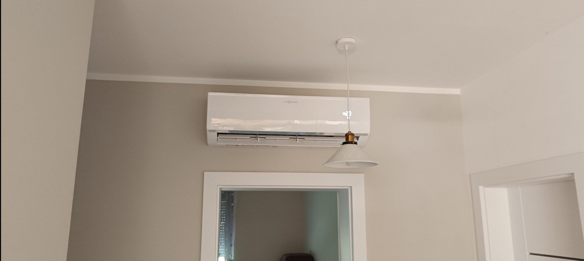 Biały klimatyzator marki Viessmann zamontowany na ścianie w jasnym pomieszczeniu, widoczny obok biały klosz lampy wiszącej i fragment ramy lustra.