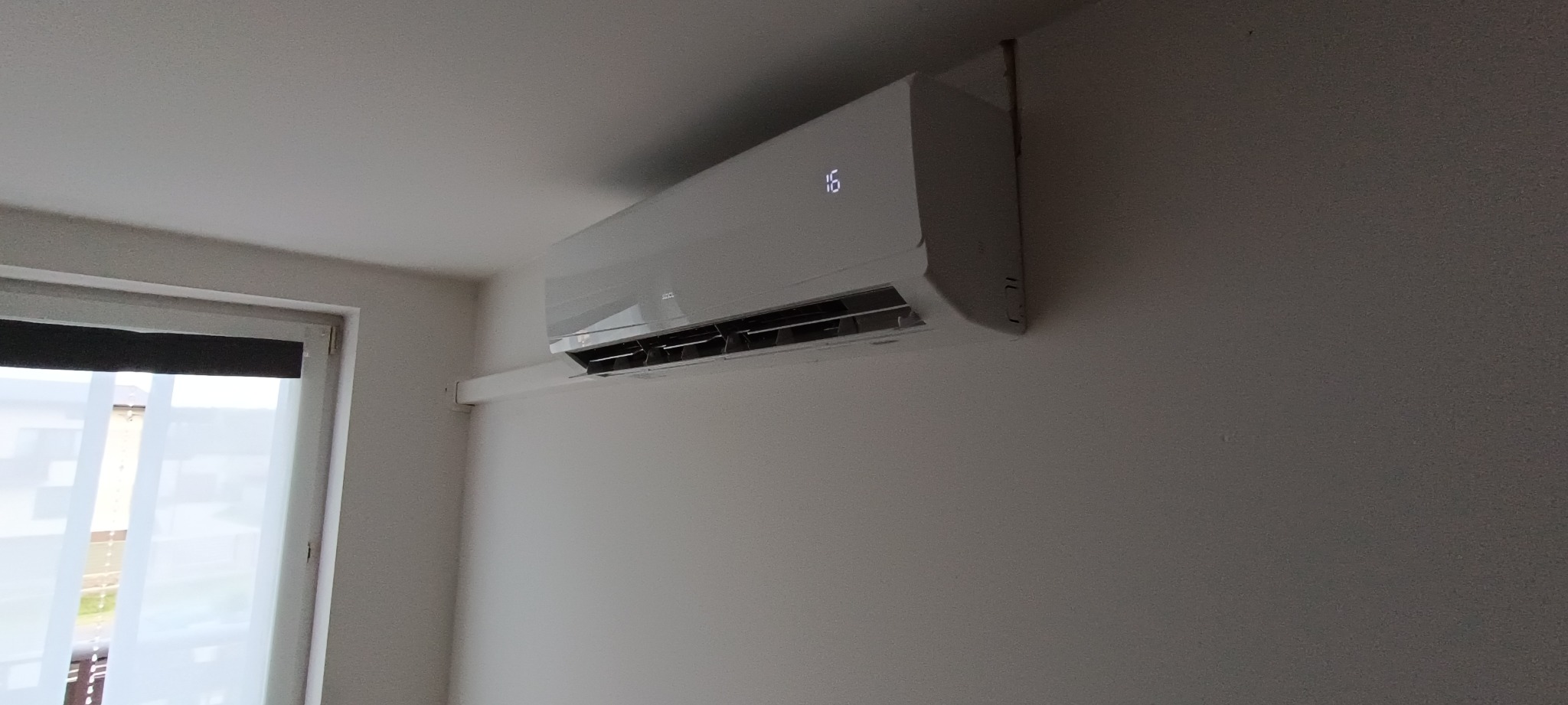 Biały klimatyzator ścienny z wyświetlaczem temperatury (16 stopni) zamontowany w jasnym pomieszczeniu, widoczny fragment okna z białymi zasłonami.