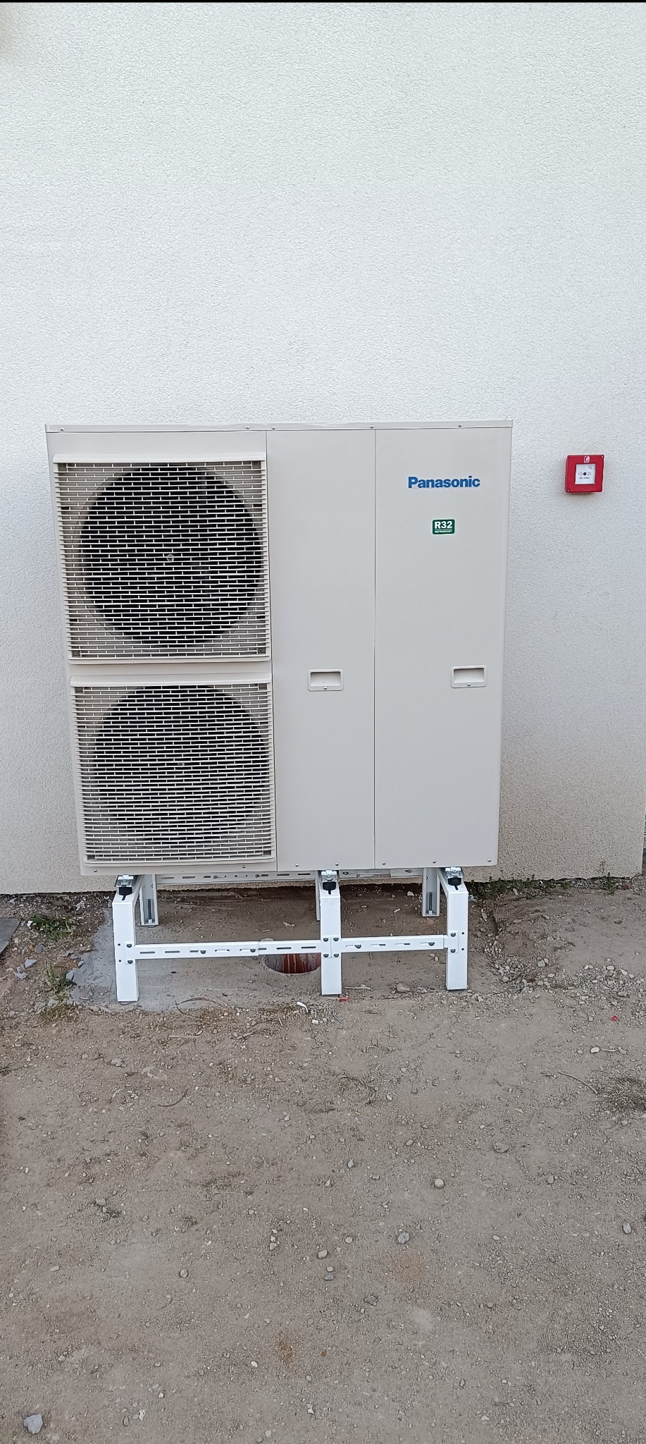 Zamontowana jednostka zewnętrzna klimatyzacji marki Panasonic z widocznym oznaczeniem R32, zainstalowana na białym stelażu przy ścianie budynku w Turku. Na ścianie widoczny czerwony przycisk alarmowy.