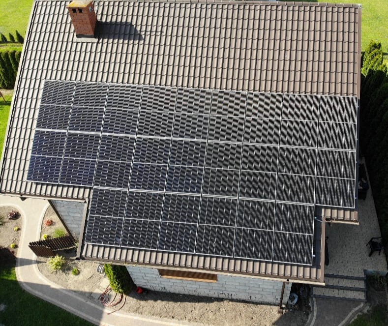 Widok z góry na dom z brązowym dachem krytym dachówką i zainstalowanymi panelami słonecznymi, obok komina. Układ paneli dopasowany do kształtu dachu, widoczne ścieżki i ogród.