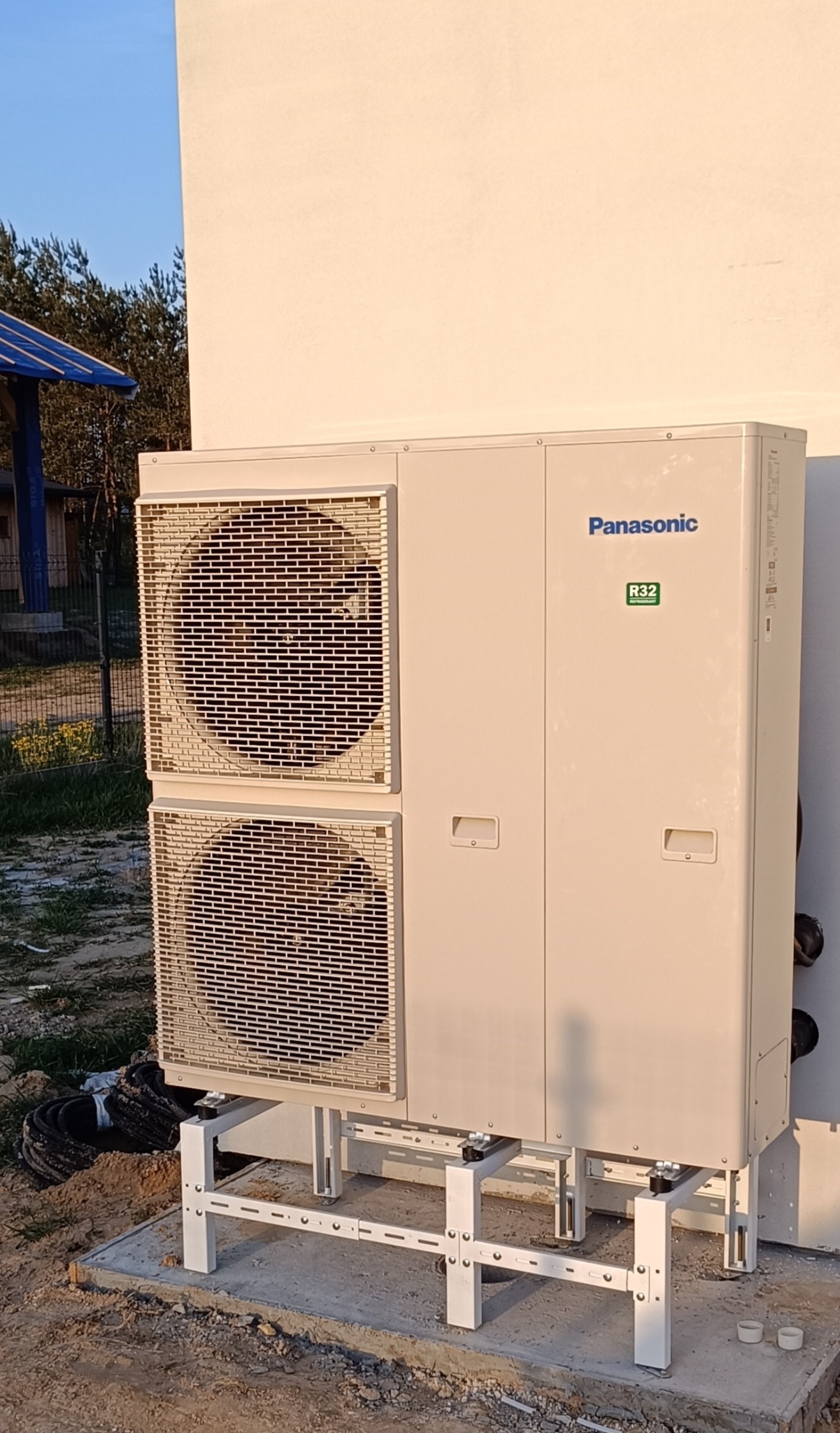 Zewnętrzna jednostka pompy ciepła Panasonic zamontowana na metalowej konstrukcji na betonowej podstawie, z widocznym fragmentem budynku w tle.