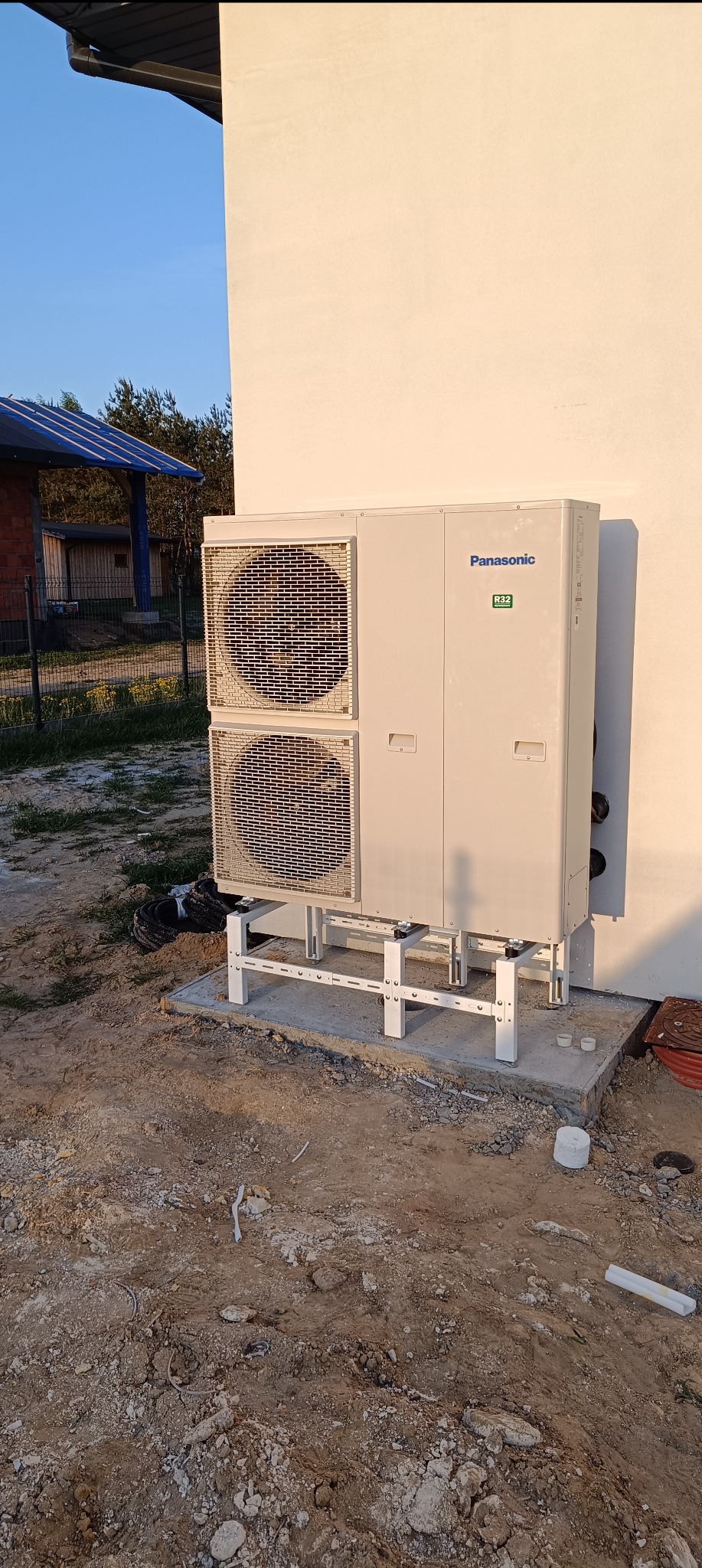 Zewnętrzna jednostka pompy ciepła Panasonic R32 zamontowana na metalowym stelażu przy ścianie budynku, widoczny fragment dachu z panelami solarnymi w tle.
