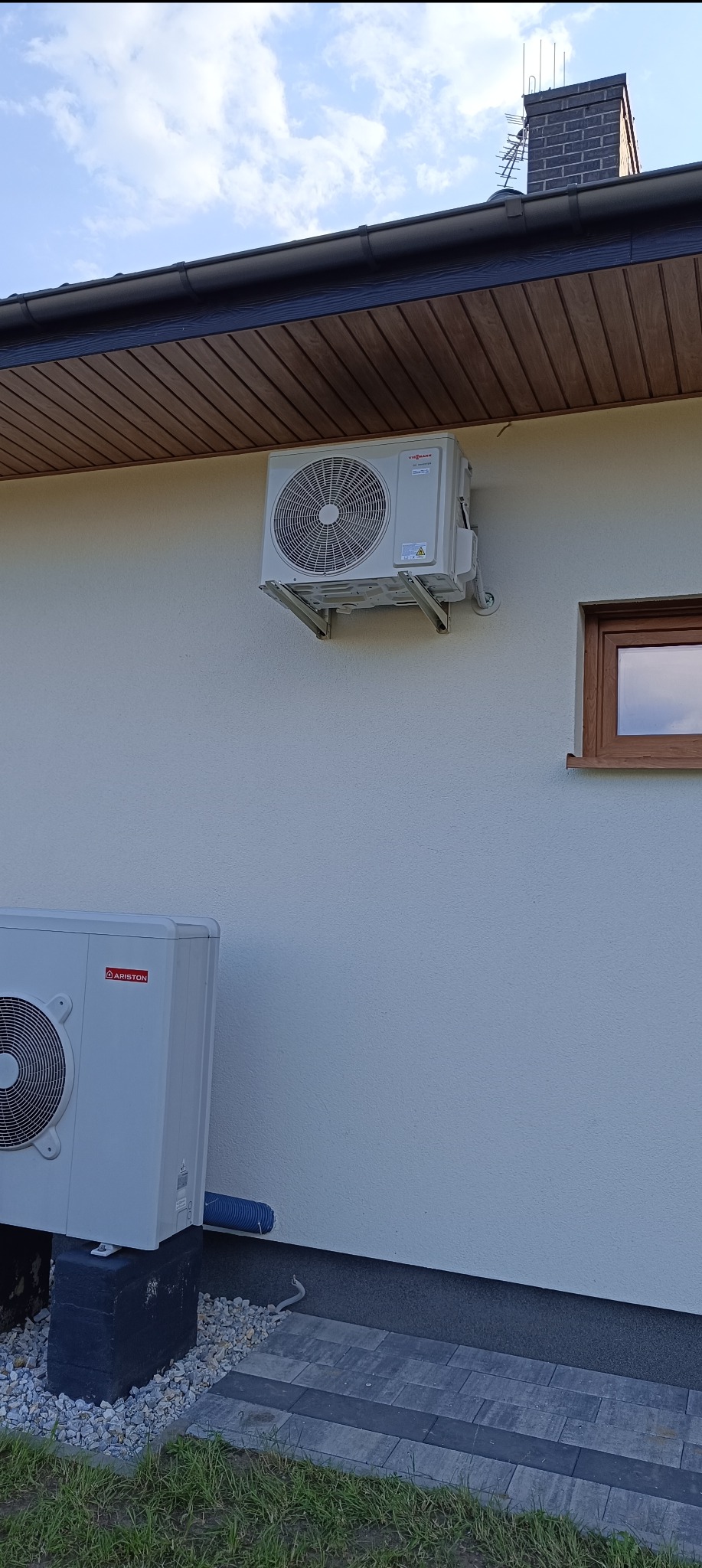 Dwa moduły klimatyzacyjne, jeden marki Viessmann zamontowany na ścianie domu pod okapem, drugi marki Ariston ustawiony na betonowym fundamencie przy chodniku z kostki brukowej.