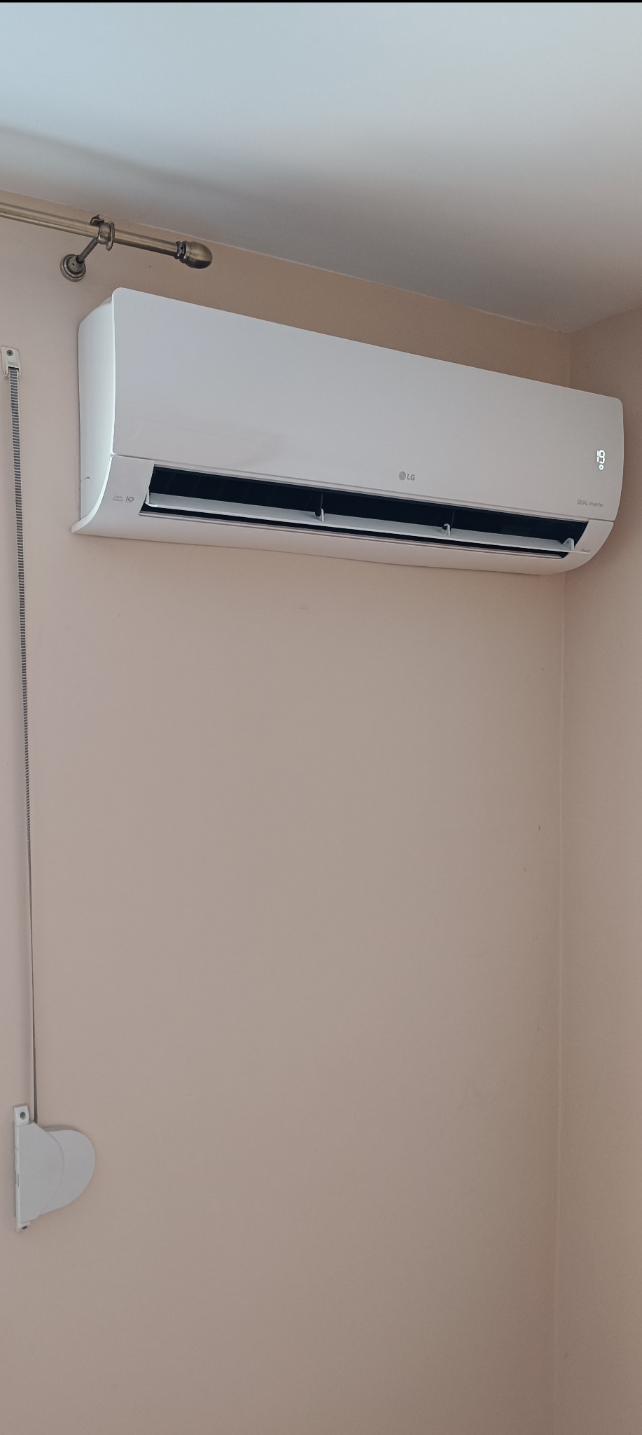Biała jednostka klimatyzacji LG Dual Inverter zamontowana na beżowej ścianie w pomieszczeniu, wyświetlająca temperaturę 19 stopni Celsjusza.