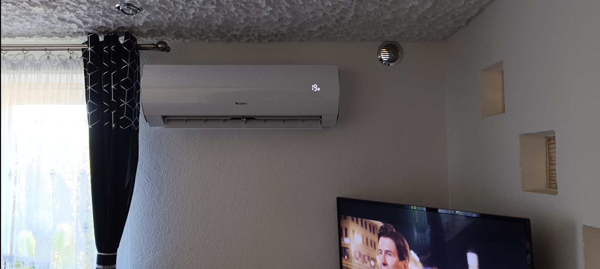 Biały klimatyzator marki Gree zamontowany na ścianie w pokoju, obok widoczny otwór wentylacyjny, telewizor i czarno-białe zasłony.