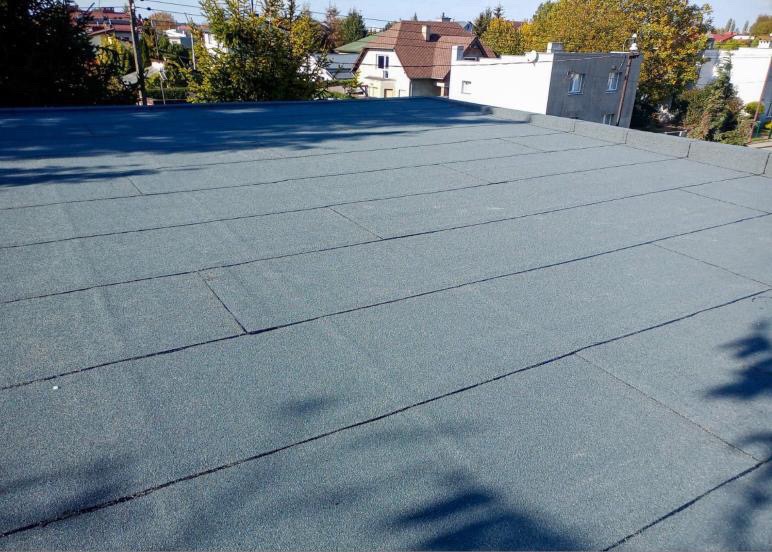 Płaski dach pokryty papą termozgrzewalną w kolorze szarym, widoczne łączenia arkuszy, w tle zabudowa jednorodzinna i drzewa.