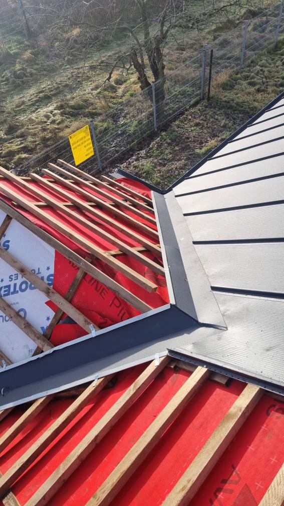 Konstrukcja dachu z łatami i membraną w kolorze czerwonym, częściowo pokryta blachą na terenie Swarzędza. Widoczne obróbki blacharskie i zabezpieczenia. W tle ogrodzenie i drzewa.