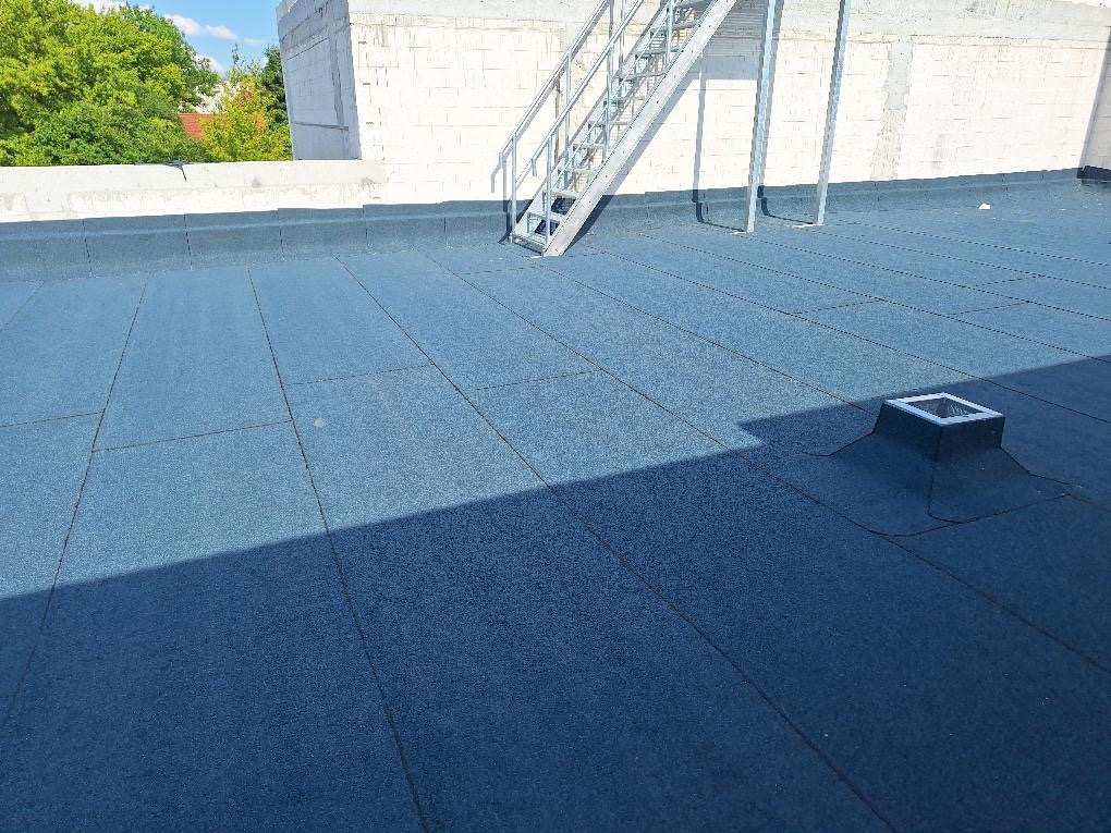 Płaski dach pokryty niebieską papą termozgrzewalną, widoczny kominek wentylacyjny i metalowe schody ewakuacyjne przy białej ścianie budynku.