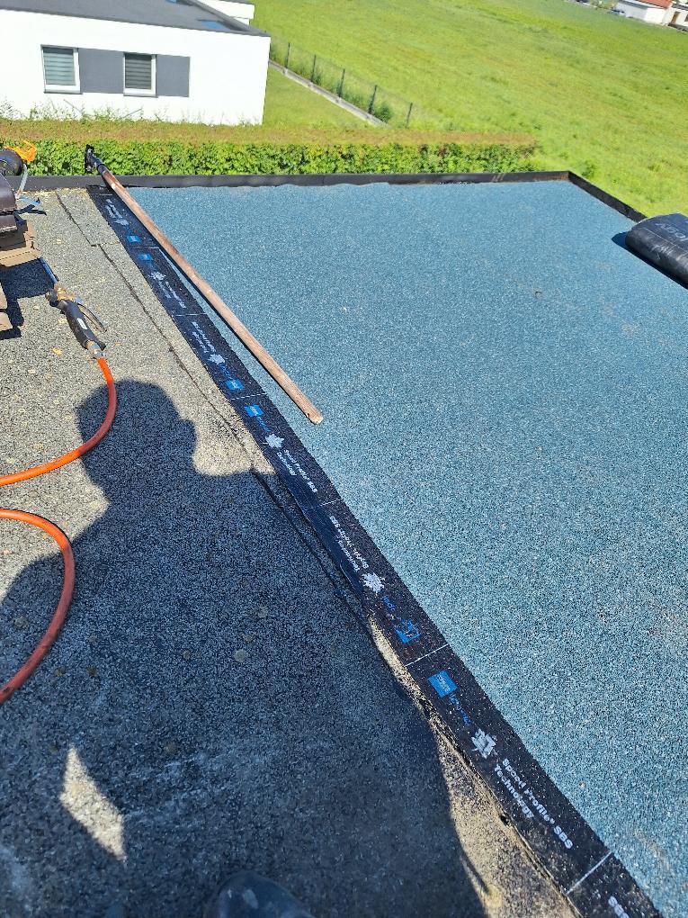 Płaski dach pokryty nową, niebieską papą termozgrzewalną, widoczne narzędzia i obróbki blacharskie w trakcie montażu, w tle budynek mieszkalny i zielone wzgórze.