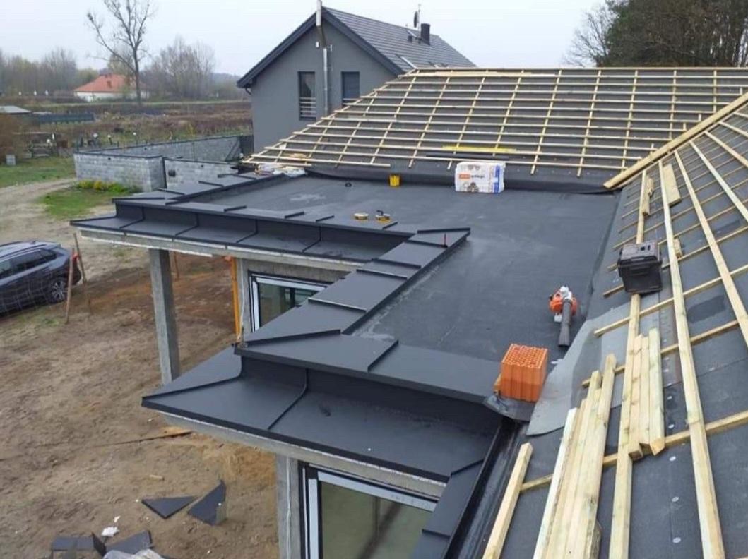 Nowoczesny dom w trakcie budowy dachu: widoczna czarna papa na płaskim dachu, świeżo zamontowane obróbki blacharskie, przygotowane drewniane łaty pod przyszłe pokrycie oraz zapas dachówek.