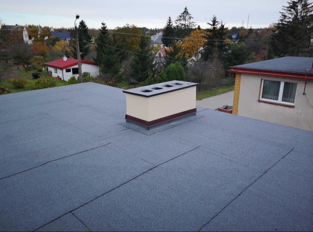 Szary dach pokryty papą termozgrzewalną, z beżowym kominem z czterema otworami wentylacyjnymi, na tle zabudowy jednorodzinnej i drzew