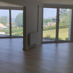 MAT-HOUSE Wykończenia Wnętrz MATEUSZ WALESIUK - Jasne, puste wnętrze z drewnianą podłogą, widokiem na zieleń przez duże okna i fragmentem schodów po lewej stronie.