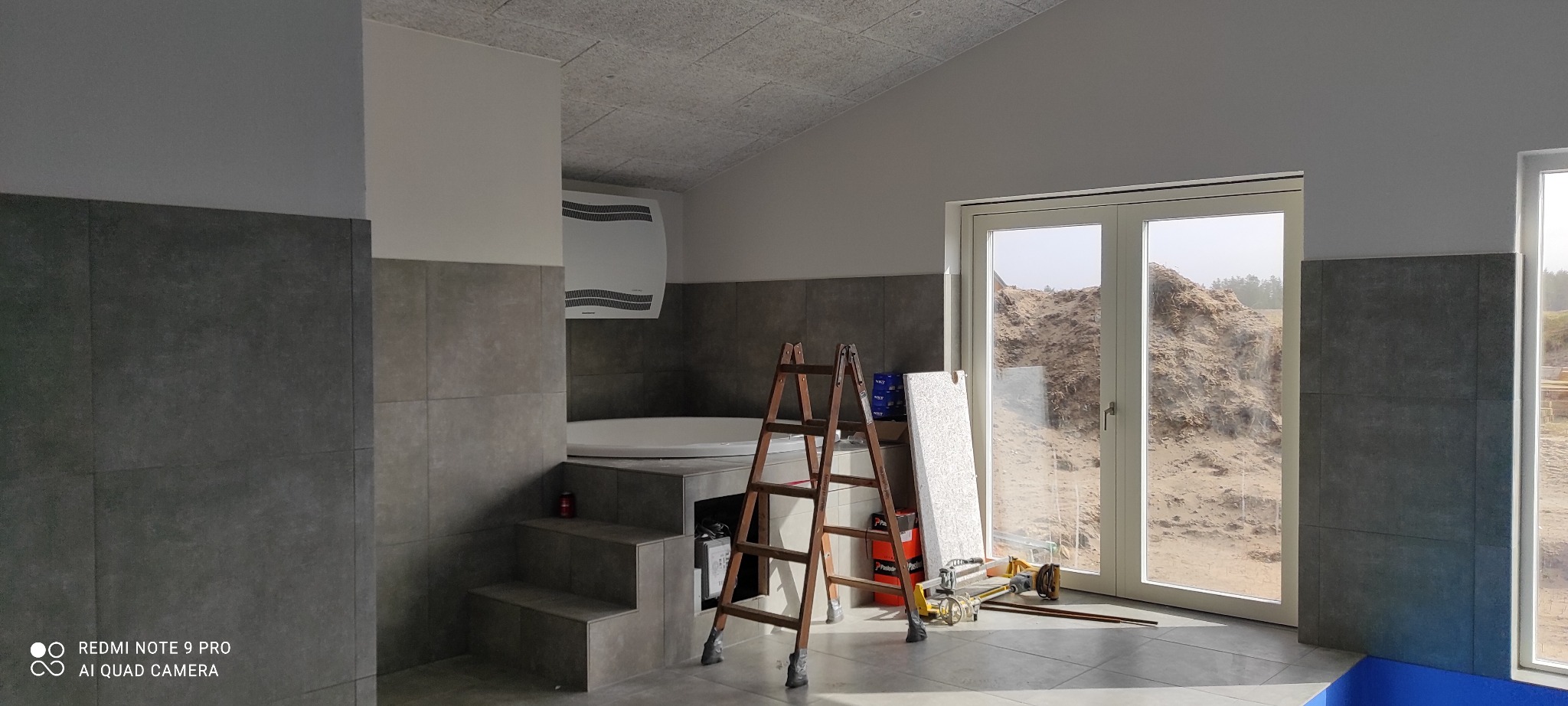 Wnętrze łazienki w trakcie prac wykończeniowych, widoczne płytki ścienne w odcieniach szarości, drabina oparta o ścianę, narzędzia oraz widok na piaszczysty teren przez okno.