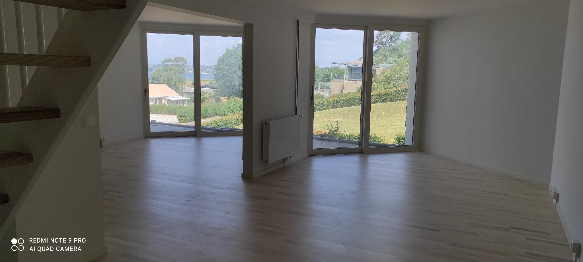 Jasne, puste wnętrze z drewnianą podłogą, widokiem na zieleń przez duże okna i fragmentem schodów po lewej stronie.