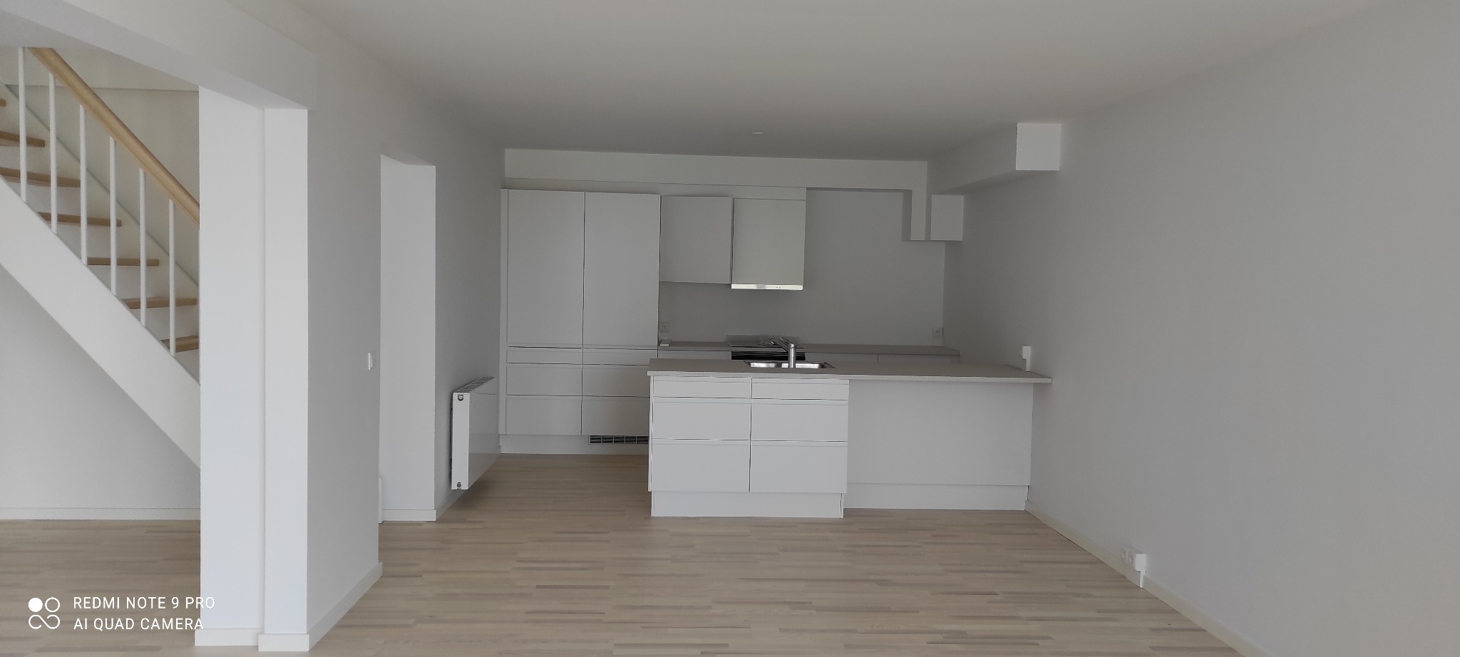 Jasne, przestronne wnętrze z minimalistyczną, białą kuchnią i drewnianymi panelami podłogowymi, widoczne schody z białymi balustradami, ściany pomalowane na jednolity, jasny kolor, grzejnik przy...